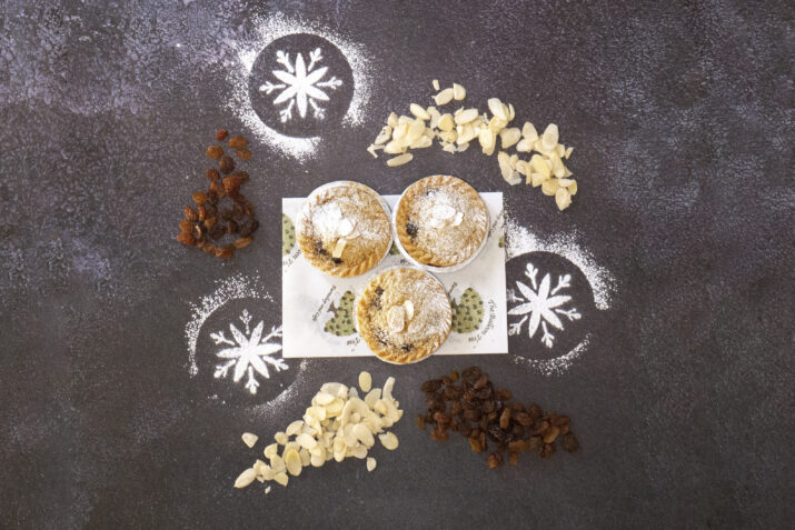 Three vegan mince pies surrounded by flaked almonds, sultanas and icing sugar snowflakes