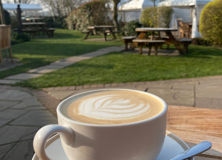 A coffee with latte art on a table with the cafe garden and trees showing behind it